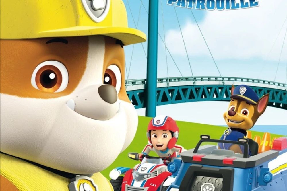 Chiot bulldog anglais portant un casque de construction jaune, souriant aux côtés de Ryder et Chase, devant un pont bleu sur fond de ciel clair.